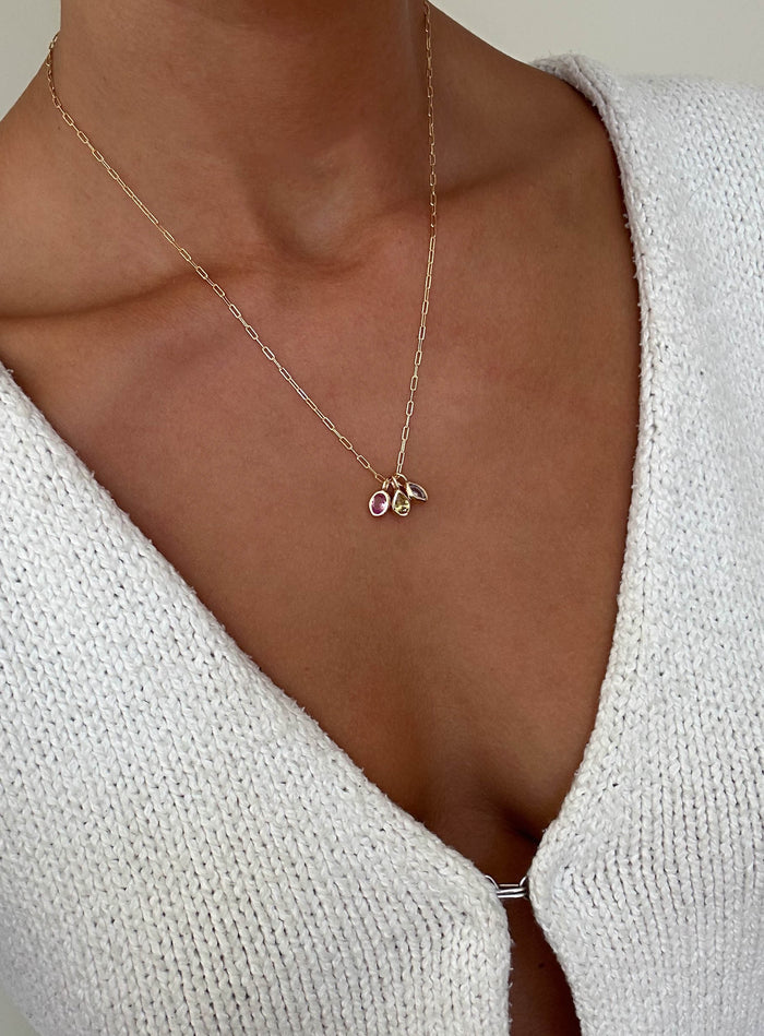A person in a white knit top with a deep neckline wears the LLUME Jewelry Build a Birthstone Necklace, featuring gold plated charms and a pink gemstone.