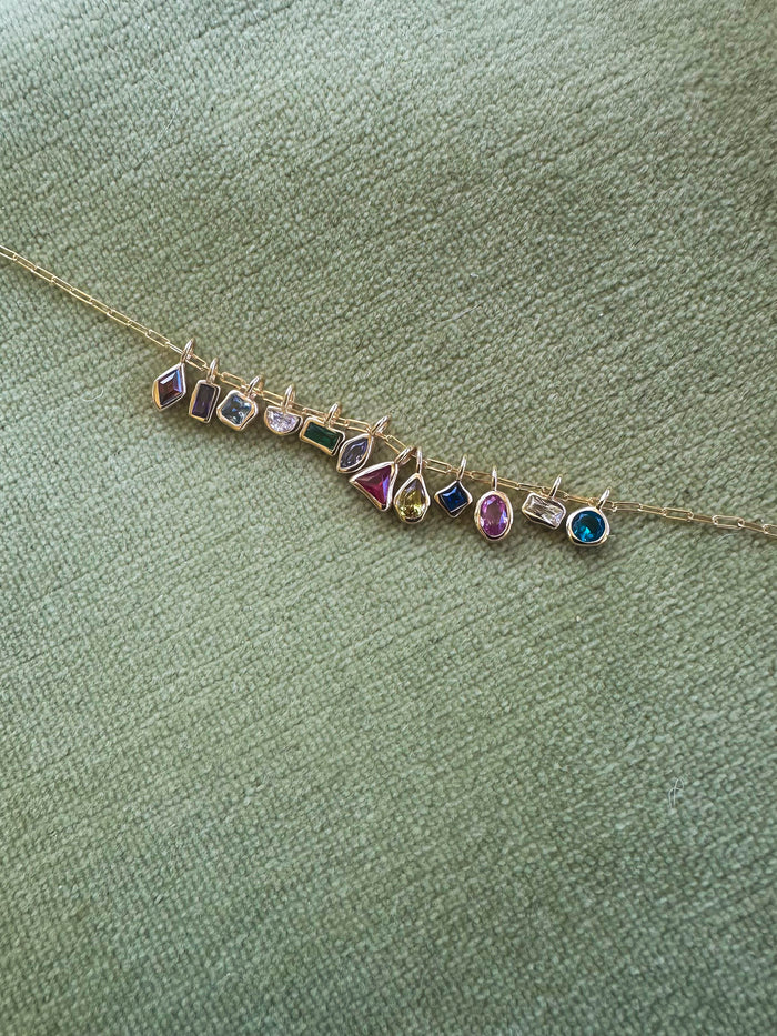 The LLUME Jewelry Build a Birthstone Necklace features gold plated charms and colorful gemstones in unique shapes, set against a textured light green fabric background.