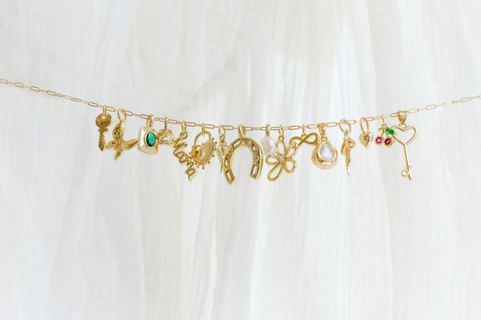 The Build a Charm Necklace by LLUME Jewelry features a gold chain adorned with gold filled charms—key, horseshoe, flower, heart, and birds—against a white fabric background.
