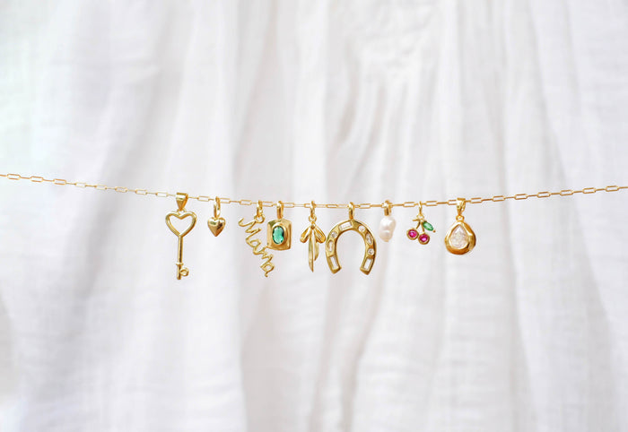LLUME Jewelry’s Build a Charm Necklace features a gold chain with nine gold-filled charms—key, heart, “mama,” turquoise stone, horseshoe, pearl with leaf, cherries, and teardrop—perfect for creating a personalized necklace. Displayed on white fabric.