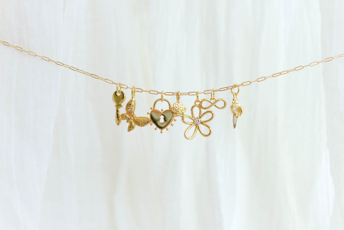 The Build a Charm Necklace by LLUME Jewelry features six unique gold filled charms—key, leaves, heart, paw print, gemmed flower, and ornate pendant—on a gold chain, elegantly displayed on soft white fabric for a truly personalized accessory.