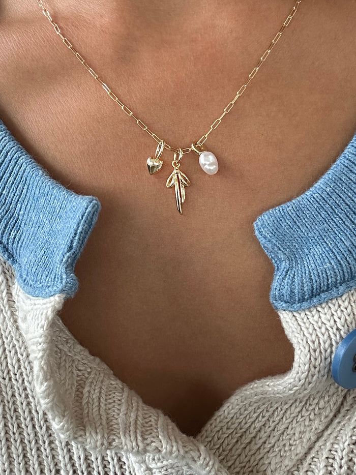 A close-up shows a person wearing LLUME Jewelry’s Build a Charm Necklace with gold filled heart, pearl, and leaf charms, styled over a cream sweater with blue collar and button for a personalized touch.