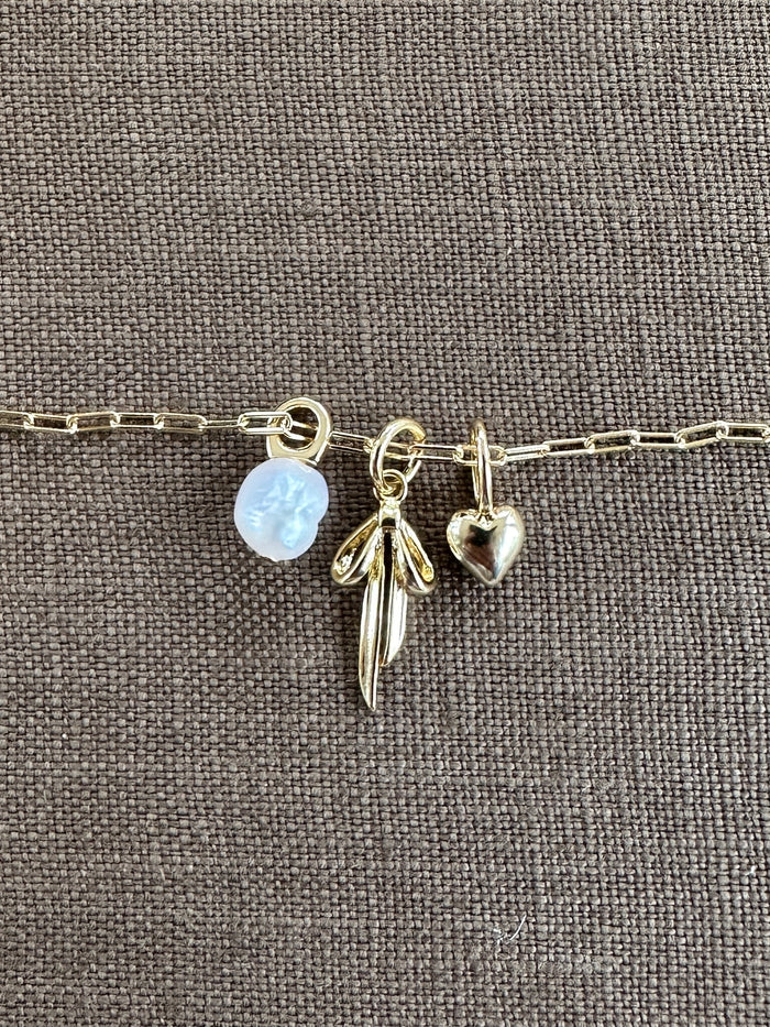 A gold chain bracelet with three gold-filled charms—a translucent flower, a wing, and a heart—from LLUME Jewelry rests on textured brown fabric, inspiring you to create your own Build a Charm Necklace or pair it with your favorite piece.