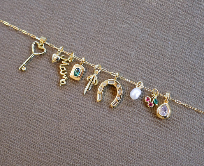 A gold chain by LLUME Jewelry showcases assorted gold filled charms—heart key, “mama” script, blue stone, and more—on a brown fabric backdrop, inspiring you to personalize your own Build a Charm Necklace.