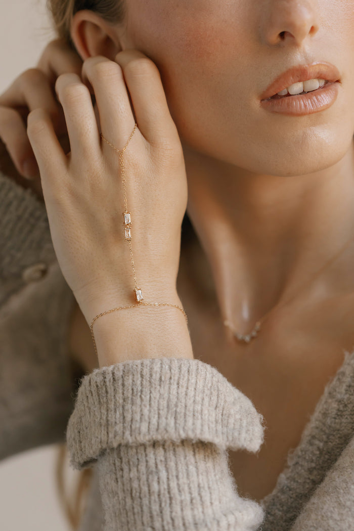 A close-up of a woman wearing the Carmen Hand Chain by LLUME Jewelry—featuring rectangular gemstones and an adjustable clasp—paired with a dainty necklace and light gray knit sweater, as she gracefully touches her face.