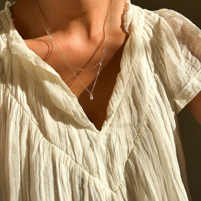 A person wears a textured off-white V-neck blouse and the LLUME Jewelry Celia Daily Set—a delicate necklace set featuring a small rectangular pendant—shown in warm natural light.