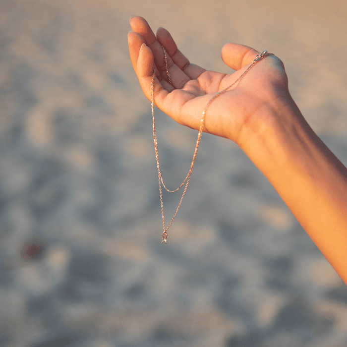 A hand is held up against a blurry sandy background, gently holding the delicate Celia Daily Set by LLUME Jewelry with its small pendant.