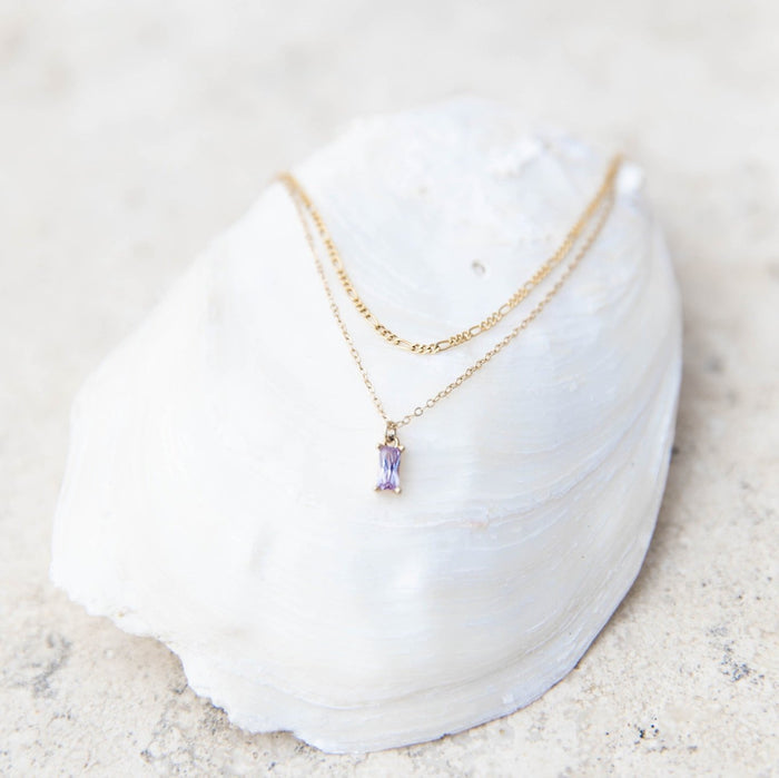 The Celia Daily Set by LLUME Jewelry, featuring a delicate gold necklace with a purple gemstone pendant, is showcased on a large white seashell atop a light sandy surface.