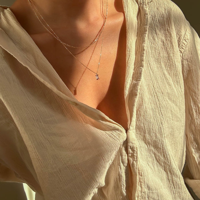 A person wears a textured cream blouse with an open collar, showcasing LLUME Jewelry's Celia Daily Set—a delicate layered gold necklace set—basking in warm sunlight.