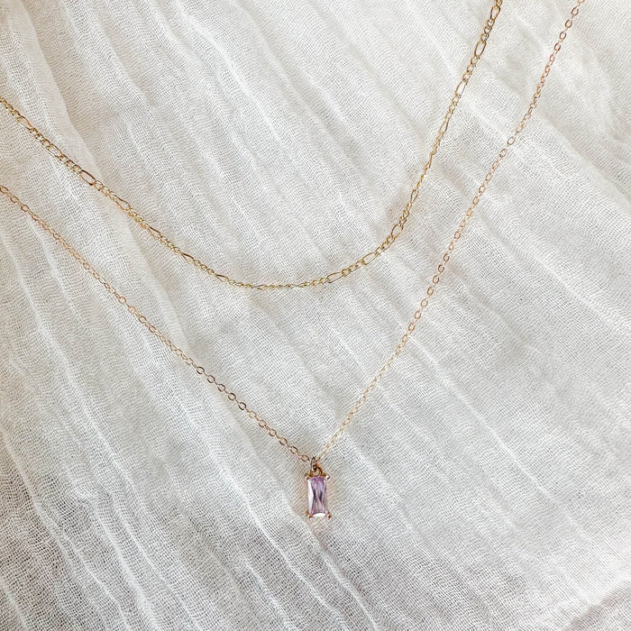 Two delicate gold chain necklaces from LLUME Jewelry's Celia Daily Set are displayed on textured white fabric—one features a light purple rectangular pendant and the other is a slightly thicker, plain chain.