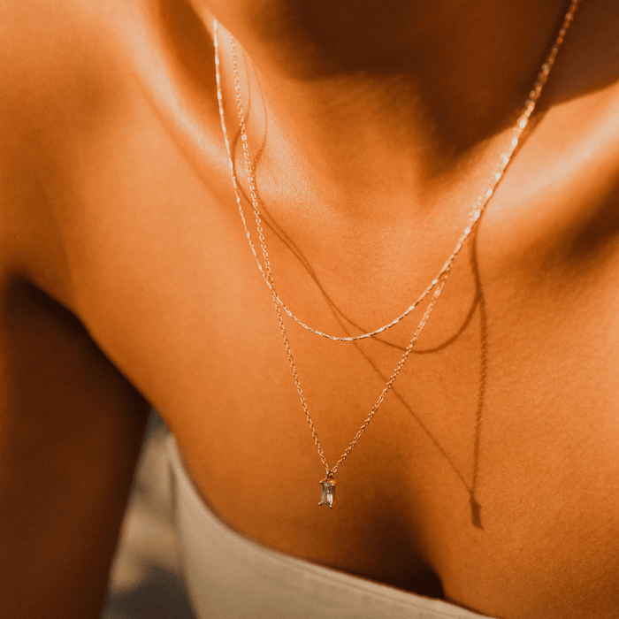 Close-up of a person wearing the LLUME Jewelry Celia Golden Set, featuring delicate layered necklaces—one with a small pendant—against sunlit skin and a light off-shoulder top.