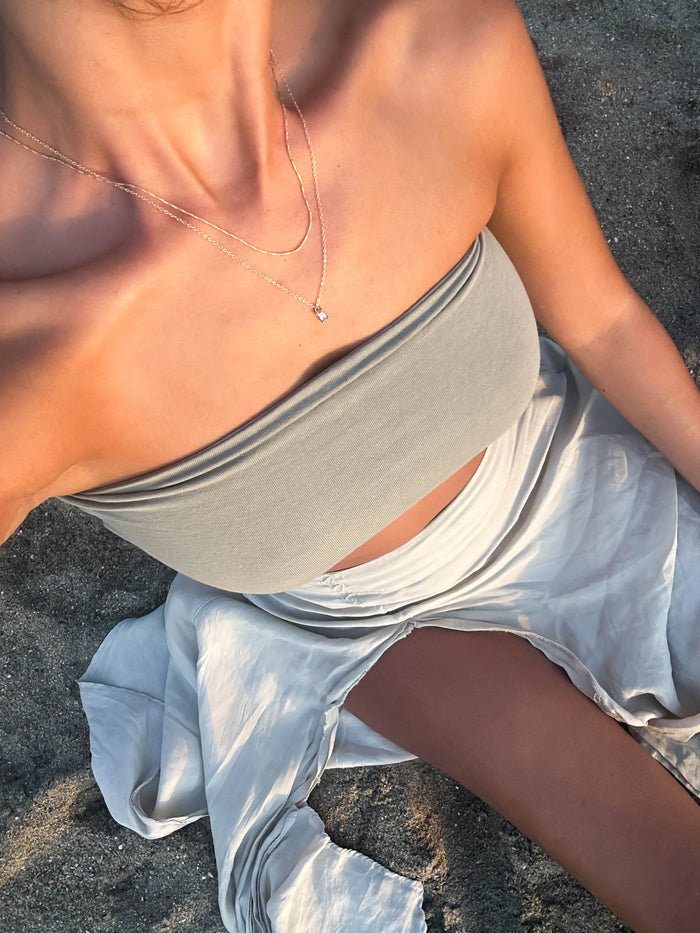 A woman sits on the sand in a light gray bandeau top, matching skirt, and the Celia Necklace from the LLUME Jewelry Celia Golden Set. Sunlight highlights her skin and outfit.