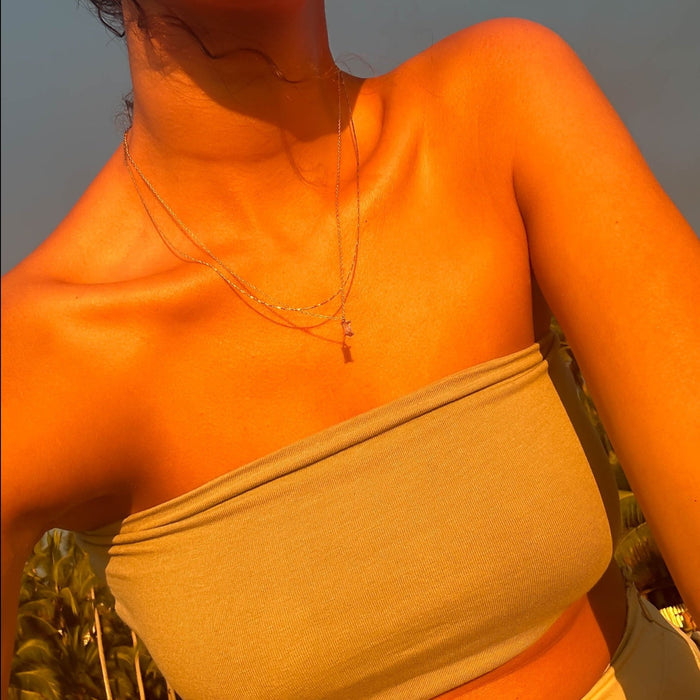 A person in a light beige strapless top wears the Celia Golden Set by LLUME Jewelry, photographed from the shoulders down in warm golden sunlight with palm leaves in the background.