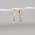 The Celia Hoops by LLUME Jewelry are gold textured hoop earrings, each with a dangling rectangular amethyst-purple gemstone, shown on a white rod against a neutral background.