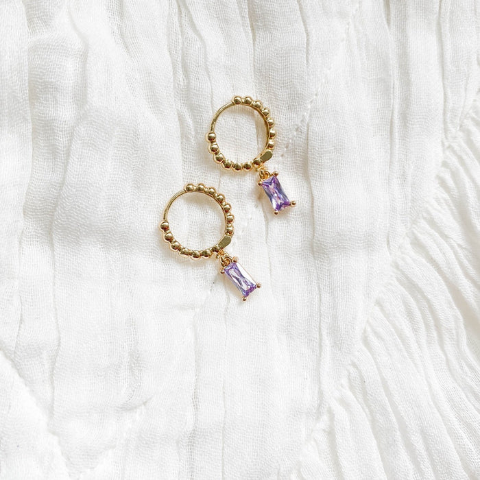 The Celia Hoops by LLUME Jewelry are gold hoop earrings with beaded detailing and a rectangular amethyst purple gemstone charm, shown on white textured fabric.