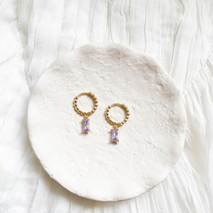 Celia Hoops by LLUME Jewelry—gold hoops with beaded details and amethyst accents, each featuring rectangular light purple gemstones, displayed on a round white dish atop pleated fabric.