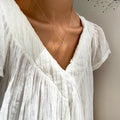 A person wears a white, pleated V-neck blouse and LLUME Jewelry’s Celia Necklace; soft light enhances the fabric’s texture and the wearer’s collarbones.