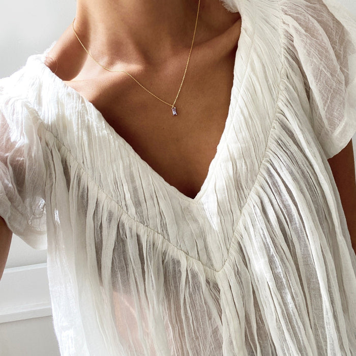 A person wears a sheer white pleated blouse with a deep V-neckline and the Celia Necklace by LLUME Jewelry in gold vermeil. The close-up image highlights the necklace’s subtle shine on the upper chest and neck.