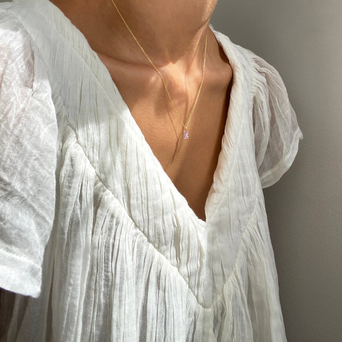 A person wearing a textured white V-neck blouse and the Celia Necklace by LLUME Jewelry—a delicate gold vermeil necklace with a small pendant—stands against a neutral background in soft lighting.