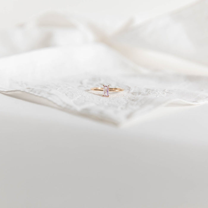 The Celia Ring by LLUME Jewelry features a delicate gold-filled band with a rectangular pale pink gemstone, displayed on a white lace handkerchief—an elegant choice for stacking jewelry lovers.