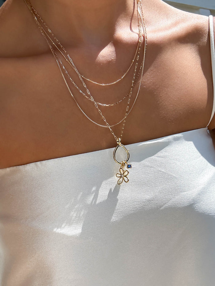 A woman wearing a white off-shoulder top and layered 18K gold filled LLUME Jewelry necklaces, including a butterfly pendant chain and the Charm Holder, in natural light.
