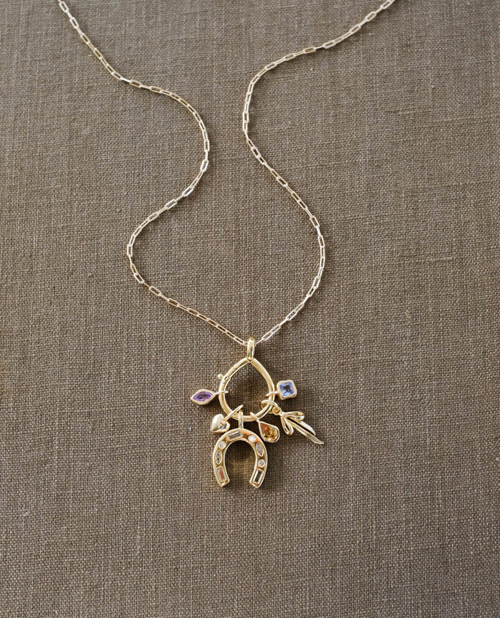 The LLUME Jewelry Charm Holder features an 18K gold-filled horseshoe pendant, small leaf and flower charms, plus two gemstone accents, all displayed on a textured brown fabric background.