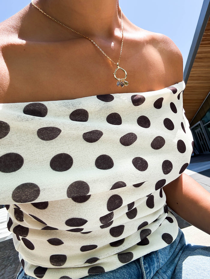 A person wearing an off-the-shoulder white top with large black polka dots, blue jeans, and the LLUME Jewelry 18K gold filled Charm Holder necklace sits outside on a sunny day with a modern building and clear sky in the background.
