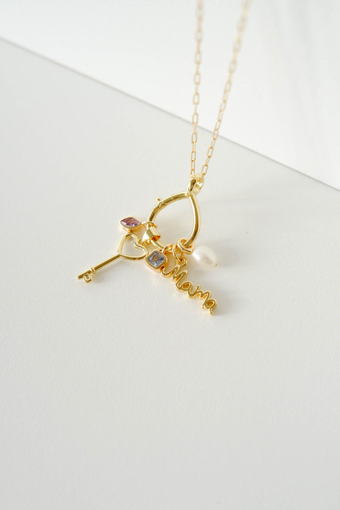 LLUME Jewelry’s Charm Holder is a chic 18K gold filled necklace with a key-shaped "Mama" pendant, pearl, and two gemstone charms—all elegantly arranged on a white surface.