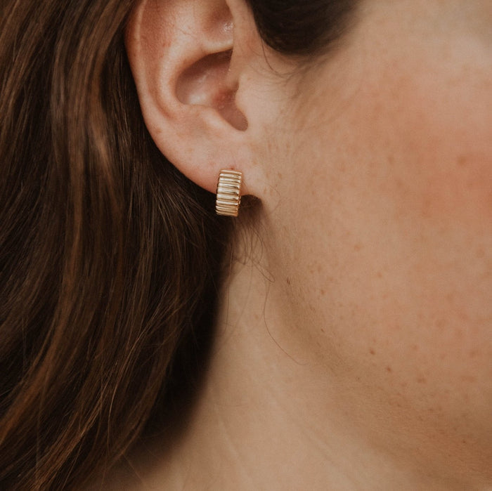 Close-up of a person’s ear adorned with the Chloe Hoops by LLUME Jewelry—small, rectangular 18k gold plated earrings with a ribbed design, radiating a subtle vintage vibe. Light freckles and brown hair highlight their timeless charm.