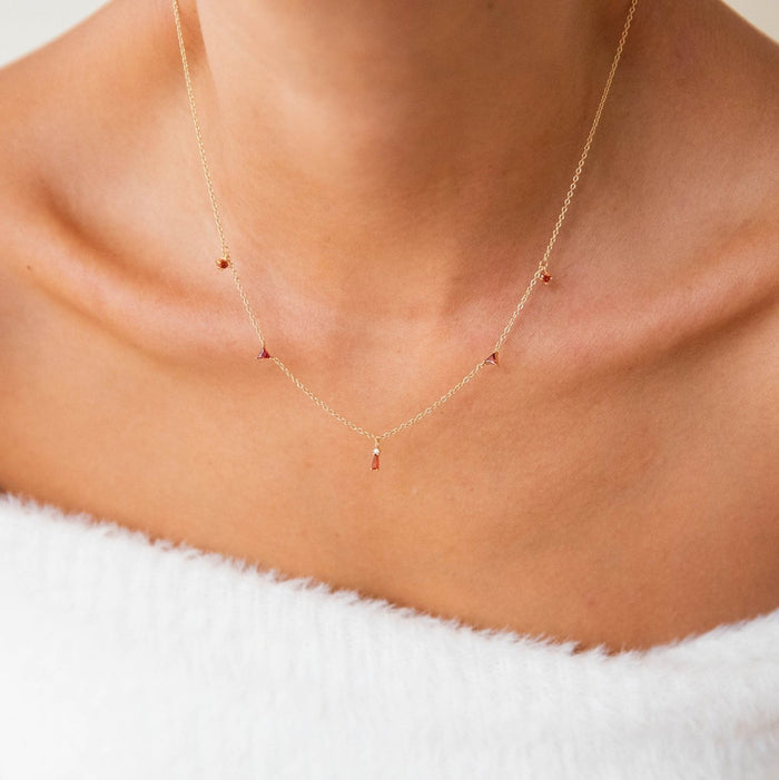 A close-up of a person wearing the Cierra Necklace by LLUME Jewelry, a gold plated piece featuring dainty ruby gemstone pendants, stands out against a white off-the-shoulder top with a neutral background for an elegant look.