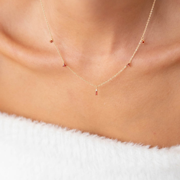 A close-up of a person wearing the Cierra Necklace by LLUME Jewelry, a dainty gold plated piece featuring small red gemstone pendants, styled against bare skin and a white, fuzzy off-shoulder garment.