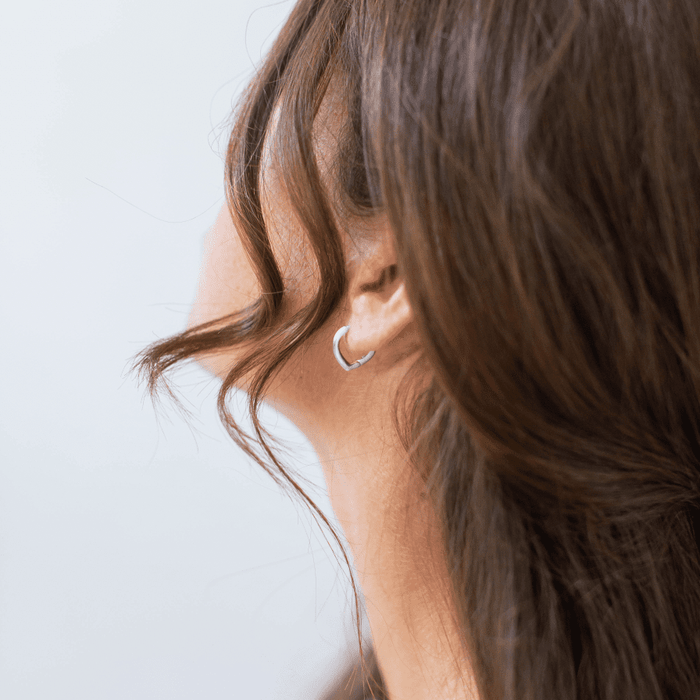 A close-up of a woman with long brown hair wears LLUME Jewelry's Classic Pointed Hoop Silver earrings in 925 silver, with a loose strand by her ear against a softly blurred background.