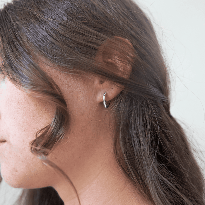 Close-up of a woman with brown wavy hair wearing the LLUME Jewelry Classic Pointed Hoop Silver earring. The small 925 silver hoop is in her ear, against a neutral, blurred background.