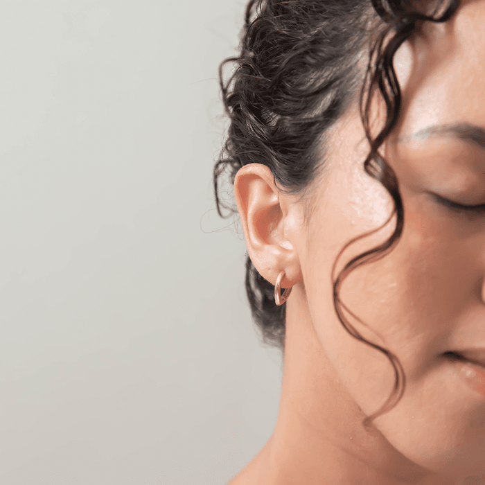 Close-up of a woman’s face with eyes closed, her ear adorned with LLUME Jewelry’s Classic Pointed Hoop earrings, curly hair framing her face against a neutral background.
