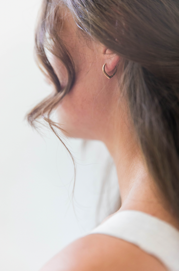 A close-up of a woman’s neck and shoulder highlights her ear adorned with LLUME Jewelry's Classic Pointed Hoop earrings. Her brown hair partly covers her face and neck, and she is dressed in a white sleeveless top.