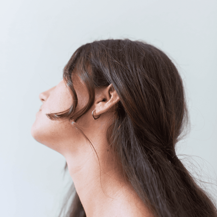 A person with long brown hair is shown in profile, facing left and wearing LLUME Jewelry's Classic Pointed Hoop earrings. A loose strand falls near their ear, set against a light, plain background.