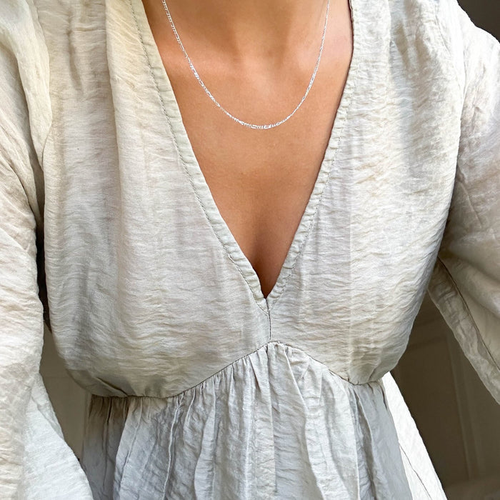 A person wears a light-colored textured blouse with a deep V-neckline, paired with the Colette Necklace Silver by LLUME Jewelry. The image is cropped to feature only the upper chest and part of the arms.