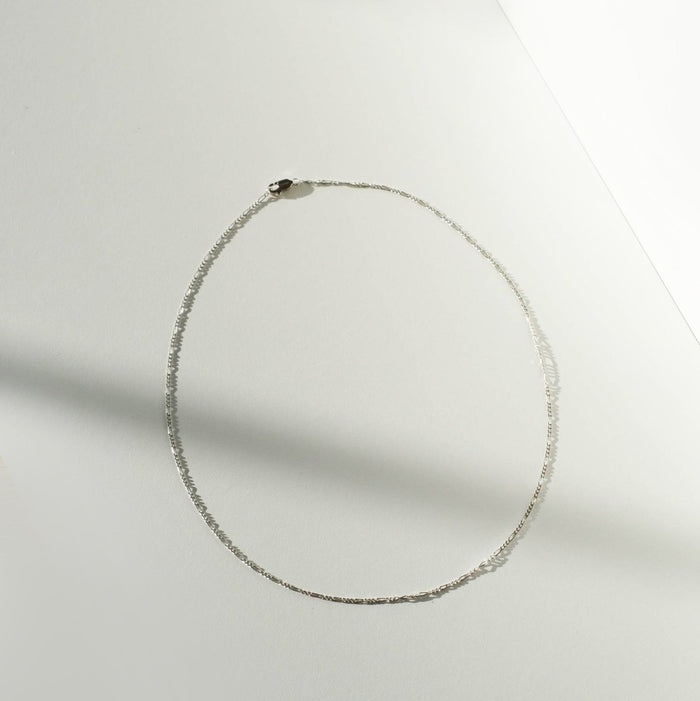 The Colette Necklace Silver by LLUME Jewelry, a delicate sterling silver chain, is arranged in an oval on a white surface with soft light and shadow creating a minimalist background.