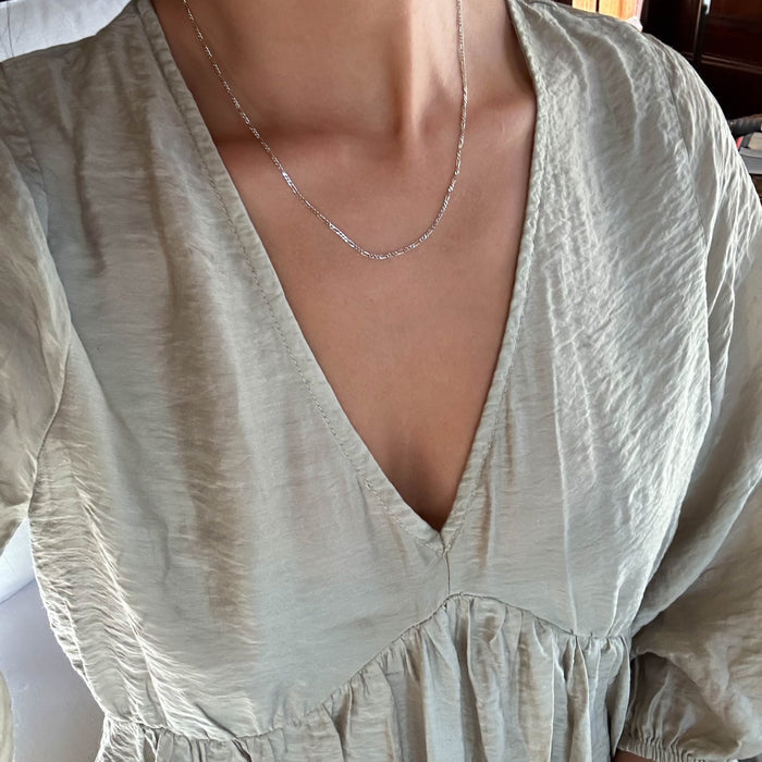 A person wearing a light-colored v-neck long-sleeve blouse is shown from the neck to upper chest, featuring the LLUME Jewelry Colette Necklace Silver—a delicate sterling silver chain.