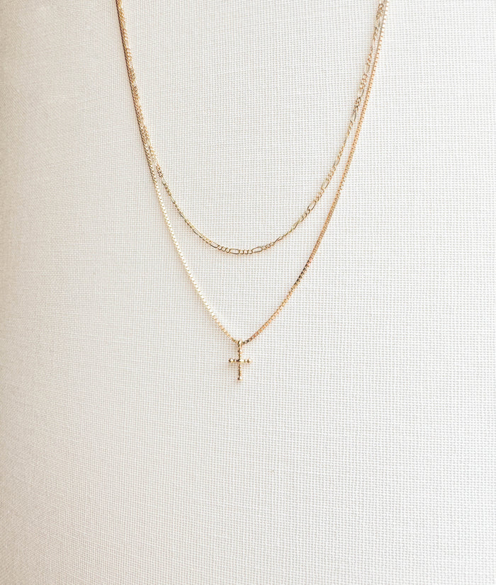 The LLUME Jewelry Cross Daily Set features three delicate gold necklaces: the Colette Necklace as the shortest, a patterned middle chain, and a classic Cross Necklace as the longest—all layered on a white textured background.