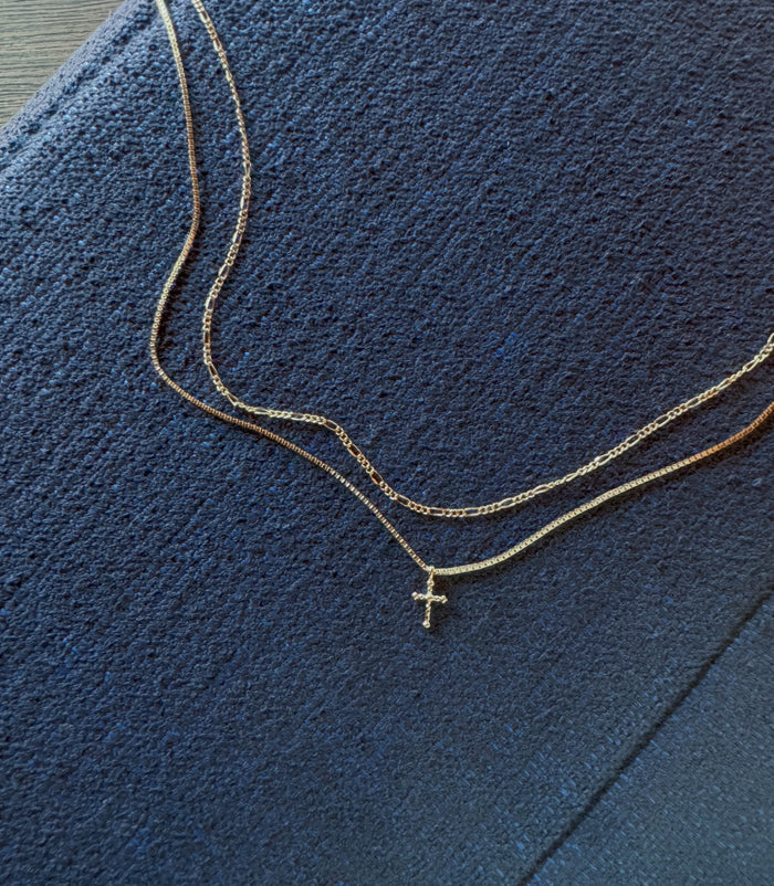 The LLUME Jewelry Cross Daily Set, featuring a Cross Necklace with a small pendant and a plain Colette Necklace, rests elegantly on textured dark fabric for a timeless look.