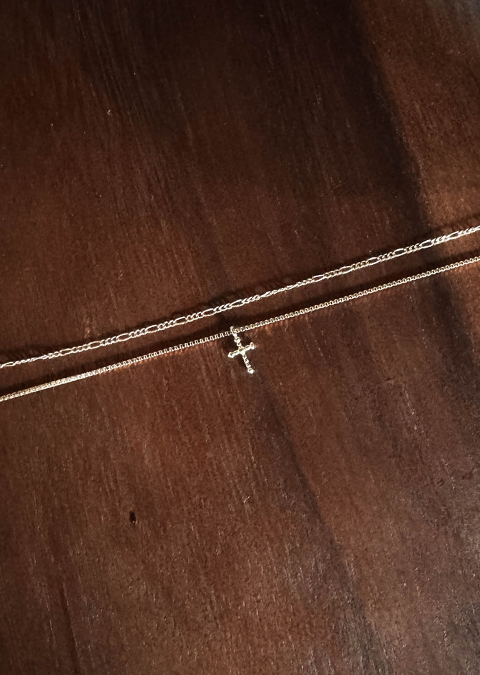 The Cross Daily Set by LLUME Jewelry features two gold chains—a minimalist cross necklace and a plain, elegant Colette necklace—displayed on dark wood, making this refined set perfect for any occasion.