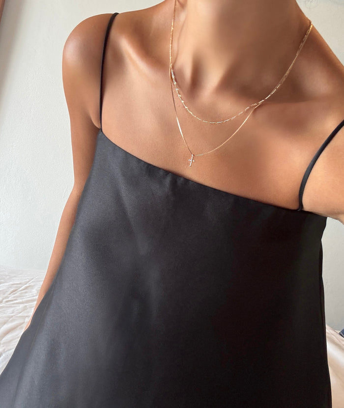 A woman poses indoors in a silky black spaghetti strap dress and the LLUME Jewelry Cross Daily Set, layered at her collarbone. The image is cropped to show only her shoulders and necklace.