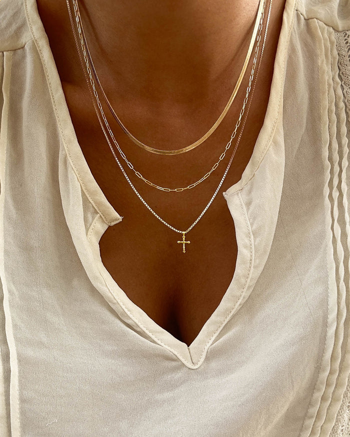 Close-up of a person wearing a cream v-neck blouse and the LLUME Jewelry Cross Necklace Set, which features a gold cross necklace paired with a delicate paperclip chain.