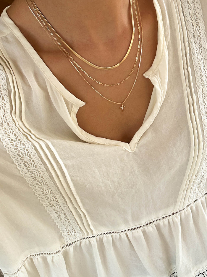 A close-up of a person wearing LLUME Jewelry’s Cross Necklace Set, featuring a layered paperclip chain and a small gold cross pendant, paired with a cream-colored lace-trimmed blouse.
