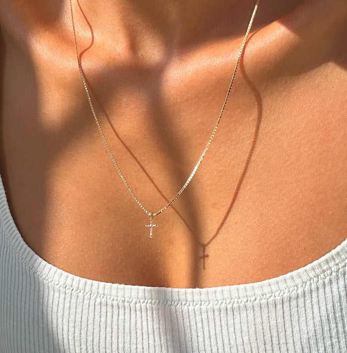 A person wears the LLUME Jewelry Cross Necklace, its delicate gold-plated pendant casting a shadow on their chest. Paired with a white ribbed tank top, bright light emphasizes both the necklace and their skin tone.