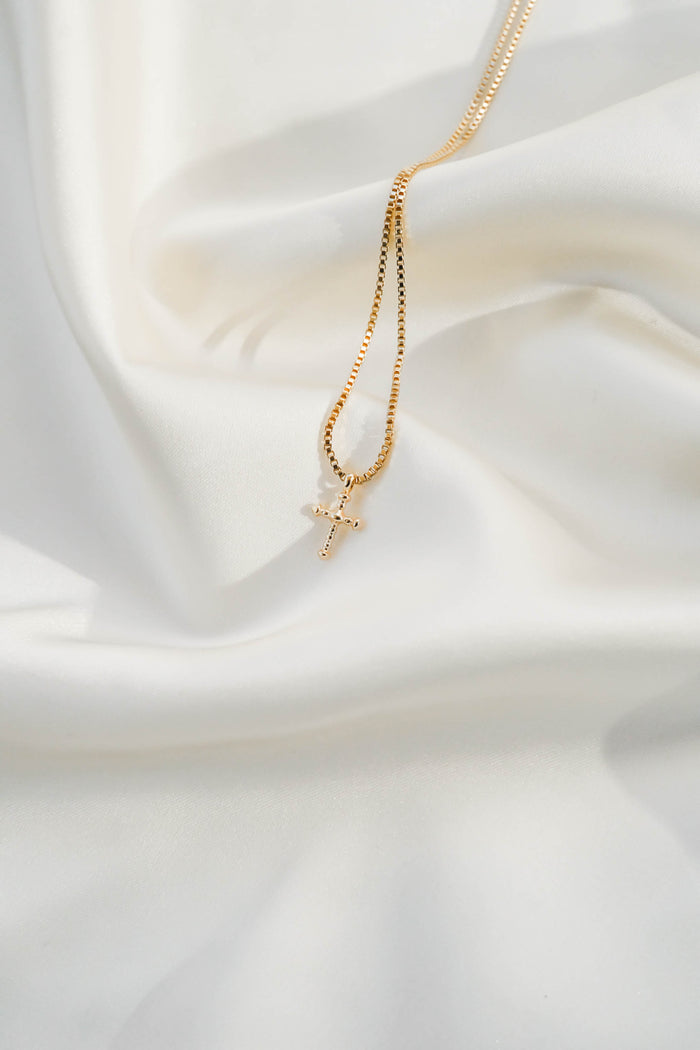 The LLUME Jewelry Cross Necklace, featuring a gold plated chain, is beautifully displayed on soft white silky fabric.