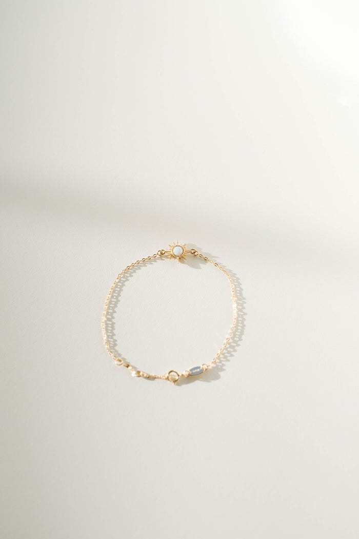 The Cyra Bracelet by LLUME Jewelry features a delicate gold-plated sterling silver band with a single light blue gemstone and a small clasp, showcased on a soft, neutral background with gentle shadows.