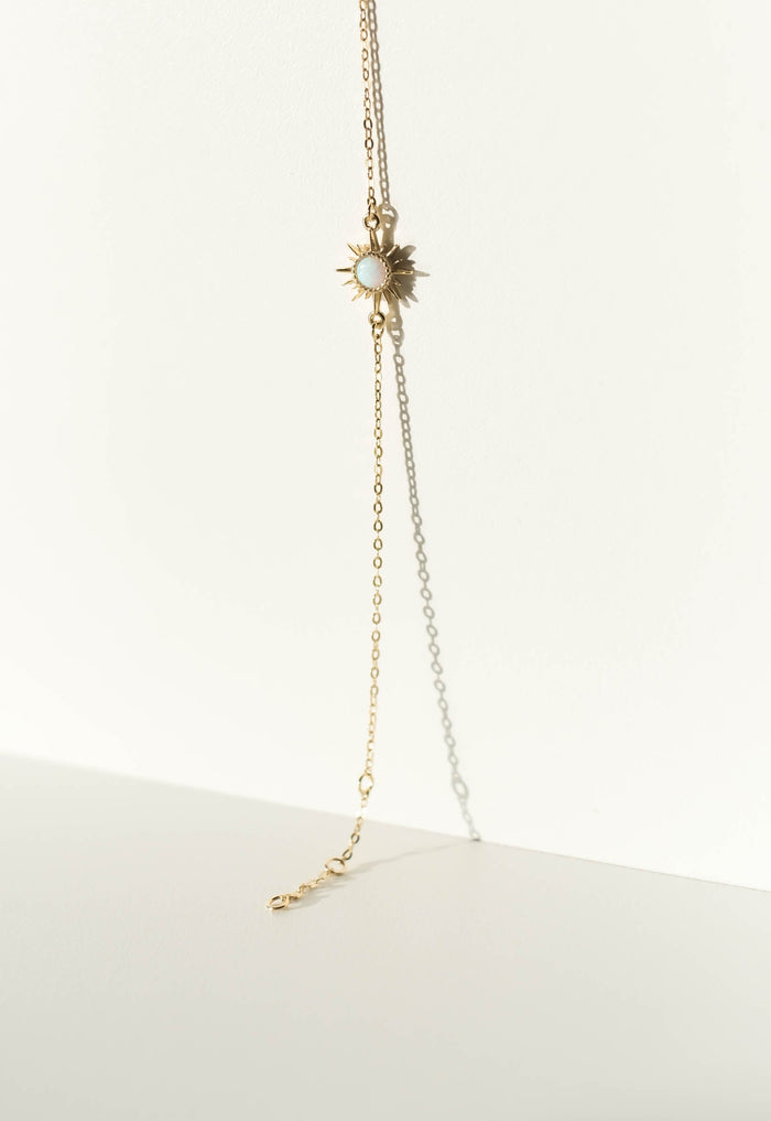 A delicate gold necklace with a sunburst pendant and white center lies on a white background, casting a soft shadow—perfectly paired with our Cyra Bracelet by LLUME Jewelry in gold plated sterling silver for a radiant look.
