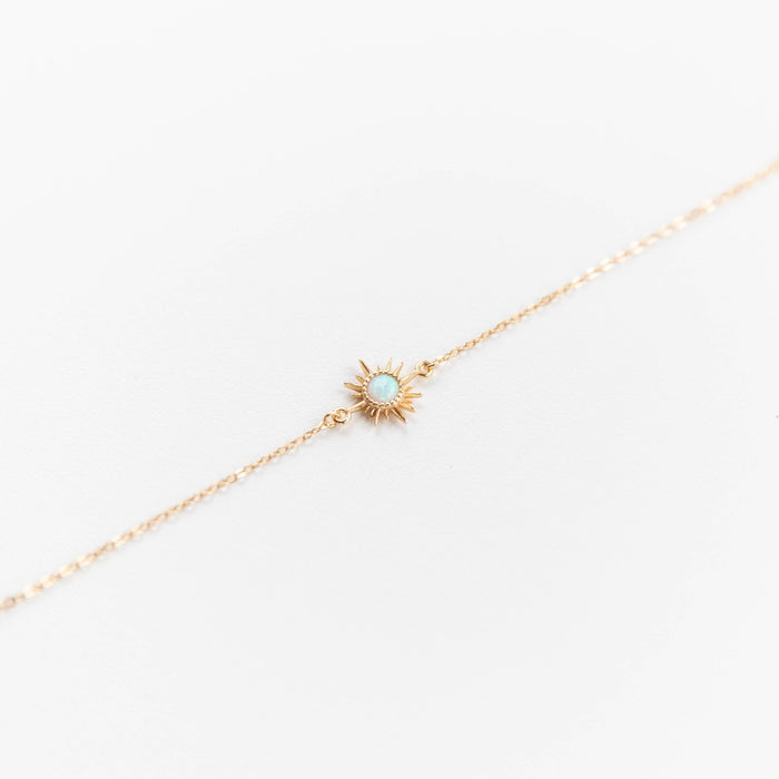 The Cyra Bracelet by LLUME Jewelry is a gold plated sterling silver piece with a tiny opal sun charm and light blue center, shown on a white background.